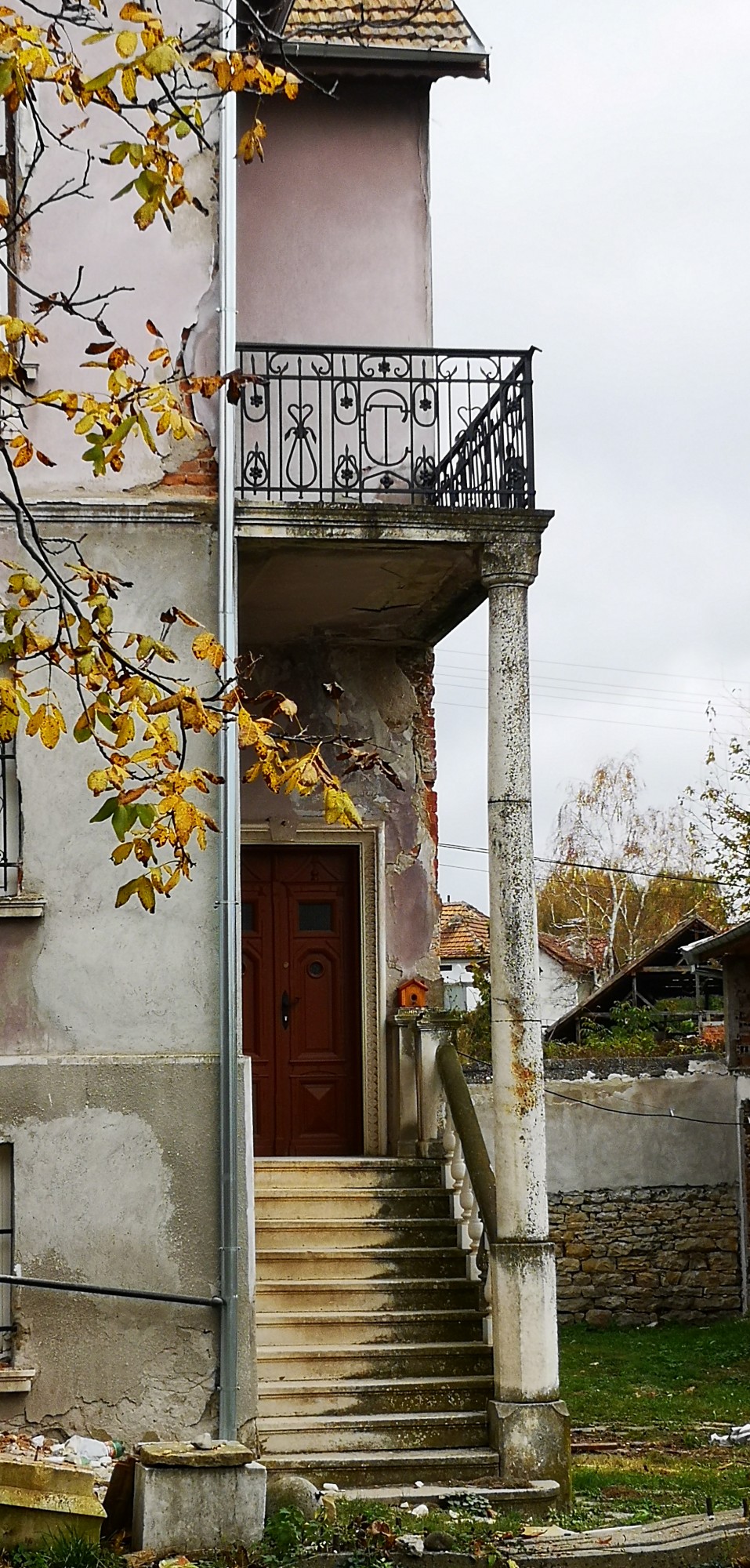 Side entrance of an old two-storey house with exterior stairs, a wooden door, balcony, metal railing, supporting column and damaged plaster.